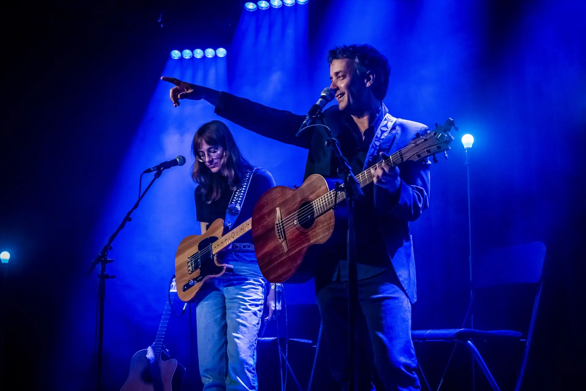 Chanteur pointant le doigt vers le haut au micro, guitariste derrière, lumière bleue.