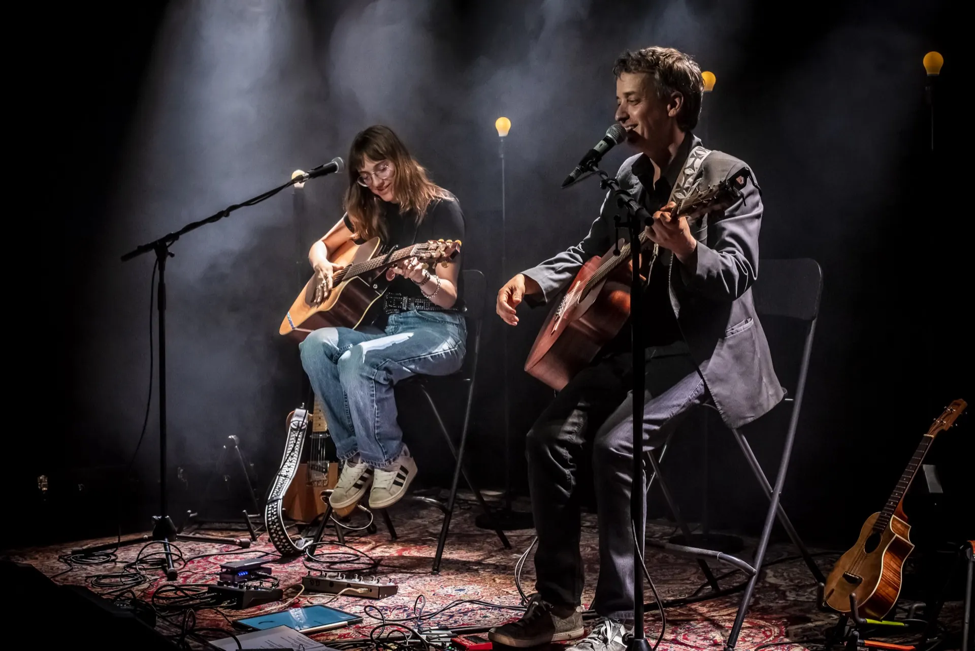 Duo assis jouant de la guitare acoustique, plan large, tapis et guirlandes lumineuses.