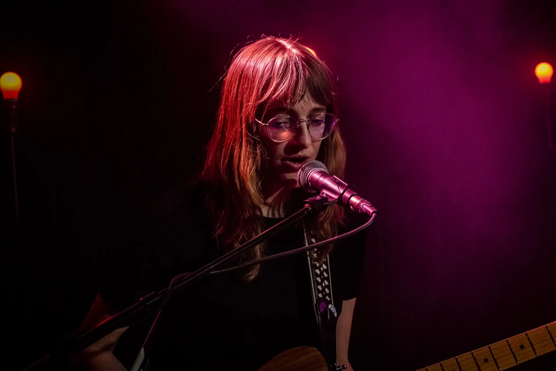 Portrait serré d'une chanteuse à lunettes au micro, fond magenta, chevelure rousse.