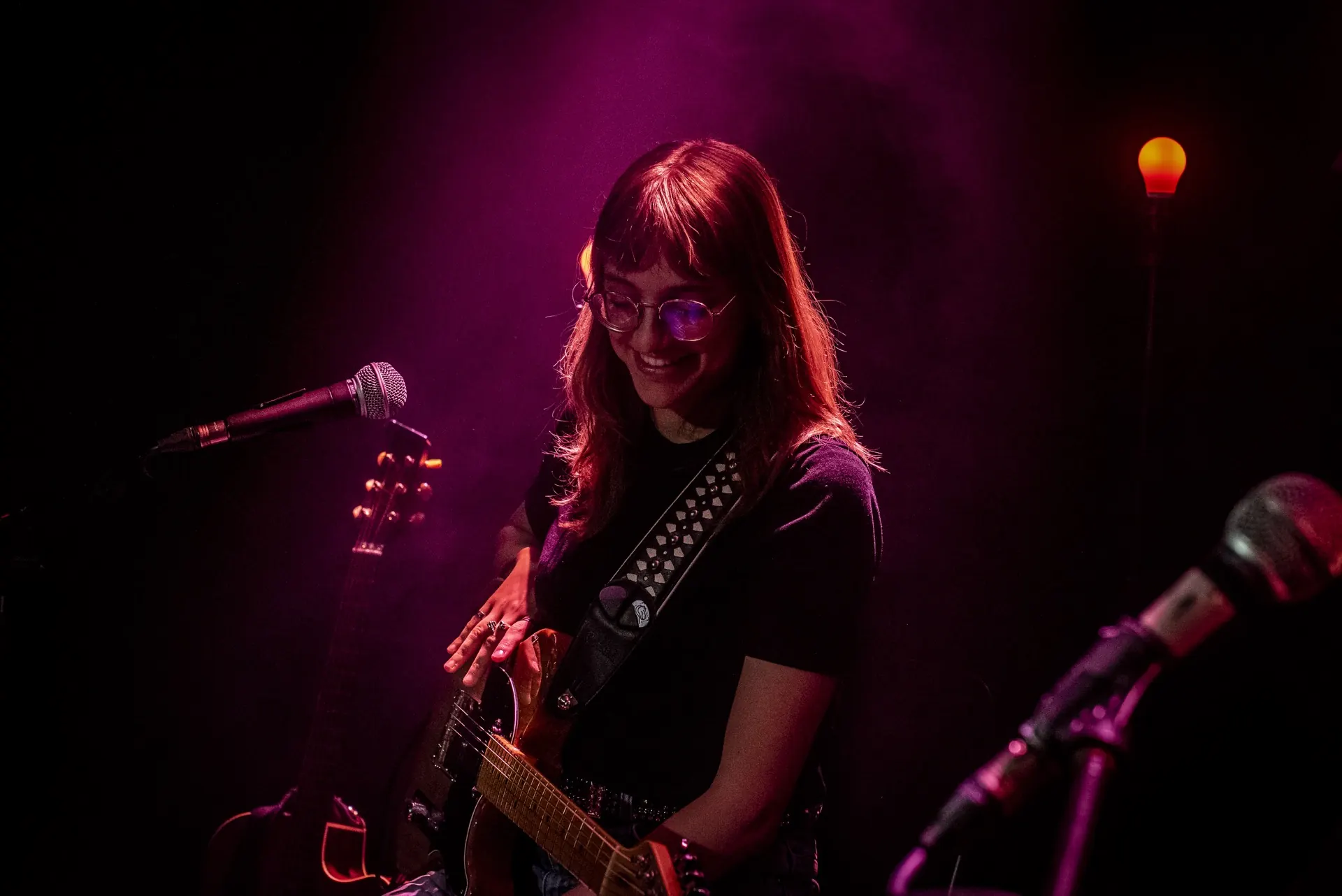 Guitariste assise souriante au micro, lunettes, guitare sur les genoux, lumière rose.