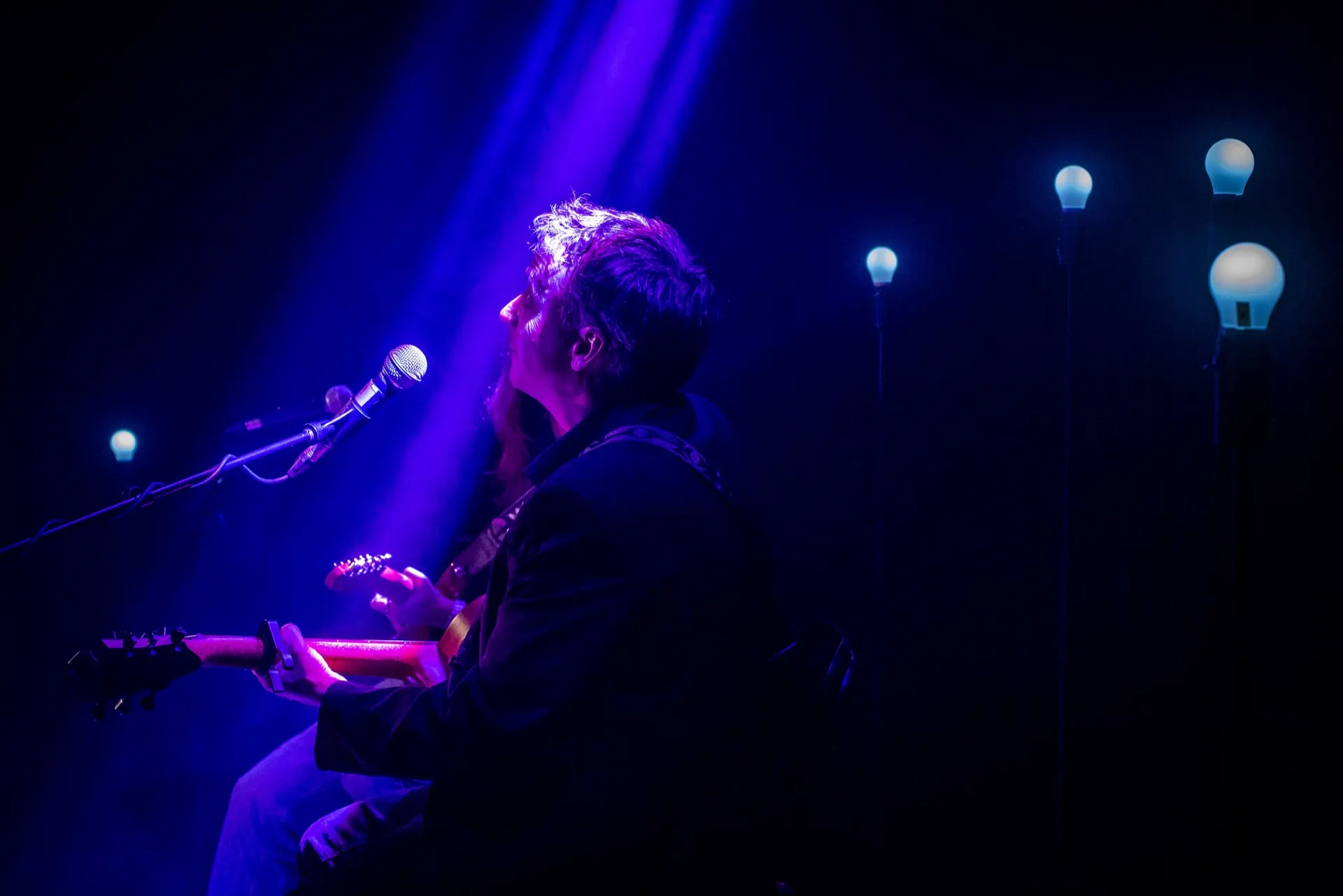 Musicien assis de profil au micro, guitare acoustique, rangée de lampes bleues à droite.
